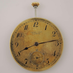 May include: A vintage pocket watch with a gold-toned face and black numerals. The watch face displays the text "HENRY CAPT GENEVE". The watch has a small seconds dial and a gold-coloured crown. The hands are black.