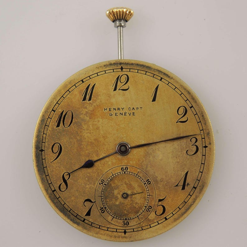 May include: A vintage pocket watch with a gold-toned face and black numerals. The watch face displays the text "HENRY CAPT GENEVE". The watch has a small seconds dial and a gold-coloured crown. The hands are black.