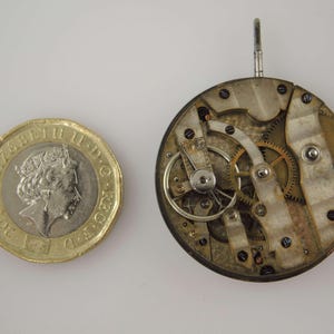 May include: A close-up of a vintage watch movement with intricate gears, levers, and screws. The watch mechanism is next to a gold-colored coin. The watch movement is round and has a silver and gold color.