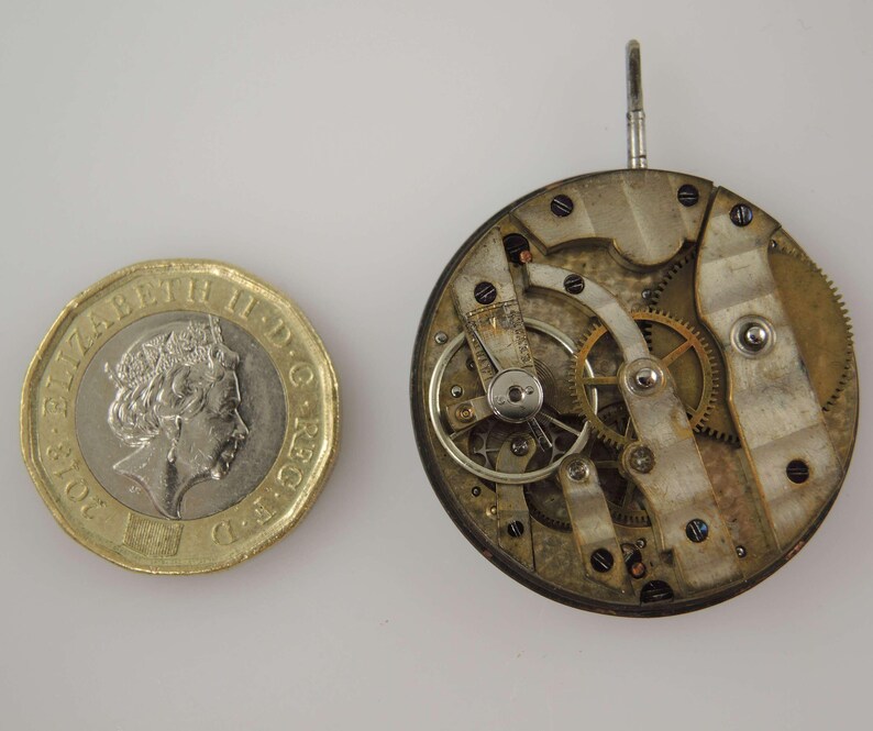 May include: A close-up of a vintage watch movement with intricate gears, levers, and screws. The watch mechanism is next to a gold-colored coin. The watch movement is round and has a silver and gold color.