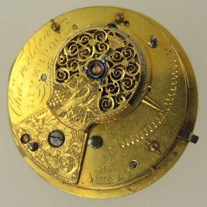 May include: Close-up of a vintage gold watch movement. The intricate mechanism features ornate filigree, engraved details, and the inscription "Chas. Fielden Liverpool." The watch's internal components are visible, showcasing its craftsmanship.