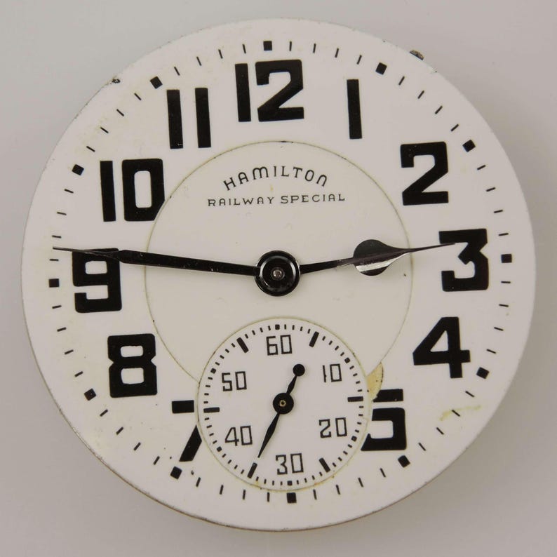 Pu&ograve; includere: Un quadrante di orologio Hamilton Railway Special vintage con sfondo bianco e numeri neri. Il quadrante include un quadrante secondario pi&ugrave; piccolo e lancette nere. Il testo "HAMILTON RAILWAY SPECIAL" &egrave; stampato sul quadrante.