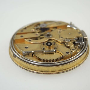 Op de afbeelding: Close-up van een gouden zakhorloge-uurwerk met ingewikkelde tandwielen en een zilveren ring aan de rand.