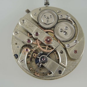 May include: Close-up of a silver pocket watch movement with intricate gears and a signed plate that reads "Swiss Made Le Coultre".