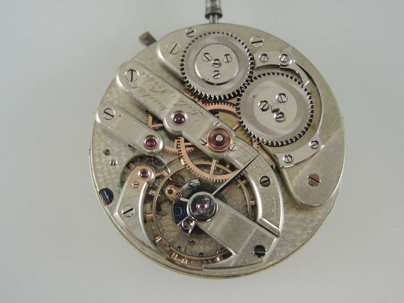 May include: Close-up of a silver pocket watch movement with intricate gears and a signed plate that reads "Swiss Made Le Coultre".