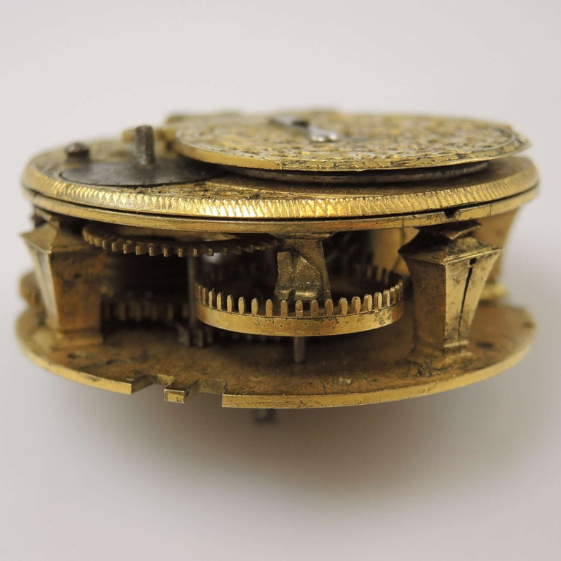 May include: A gold-toned pocket watch with intricate gears and a circular face. The watch is disassembled and the inner workings are visible.