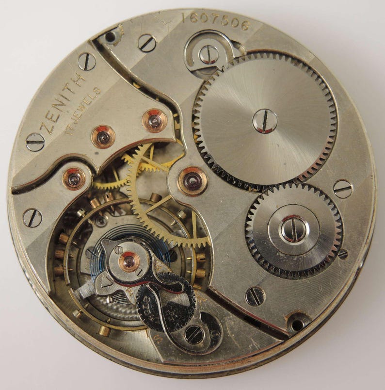 May include: Close-up of a Zenith watch movement with visible gears, screws, and other internal components. The watch movement is silver-toned with gold-colored gears and the word "ZENITH" is visible. The number "1607506" is also visible.