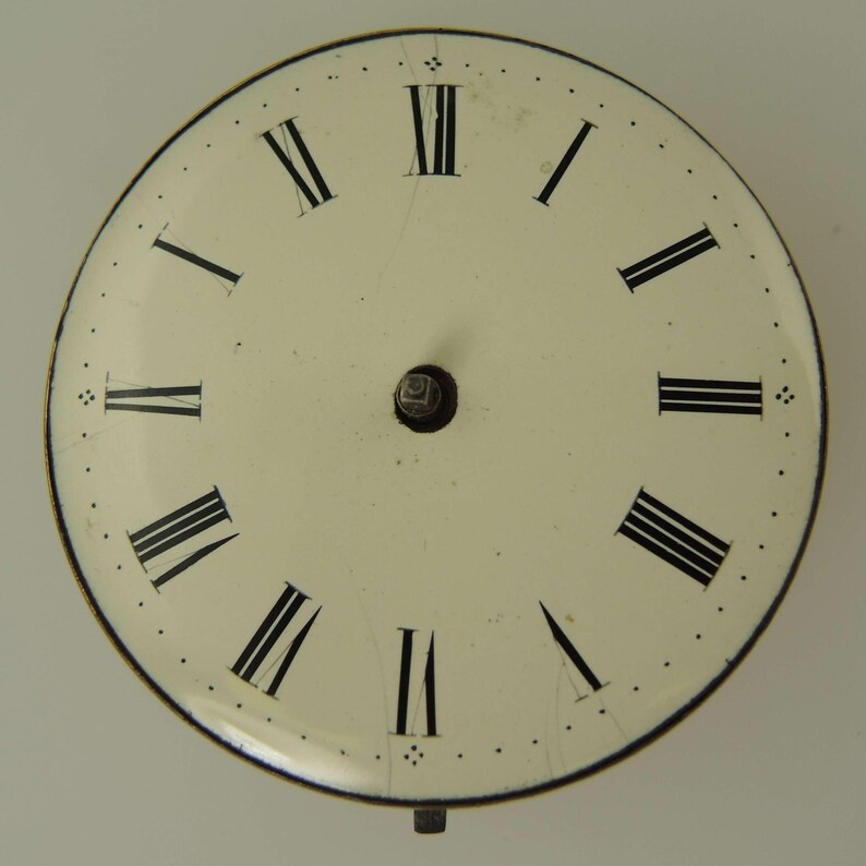 May include: Cream-colored clock face with black Roman numerals and a small central post. The face has a black outer ring and small dots marking the minutes. The clock face shows signs of age.