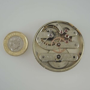 May include: A silver pocket watch movement with intricate gears and a gold-colored winding stem. The watch movement is about the size of a US one-pound coin.