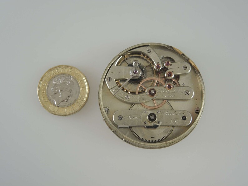 May include: A silver pocket watch movement with intricate gears and a gold-colored winding stem. The watch movement is about the size of a US one-pound coin.