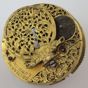 May include: A gold pocket watch with intricate floral engravings and a detailed face. The watch face has a small, round window and the text "DE CHAINE I OND ON" is visible on the bottom.