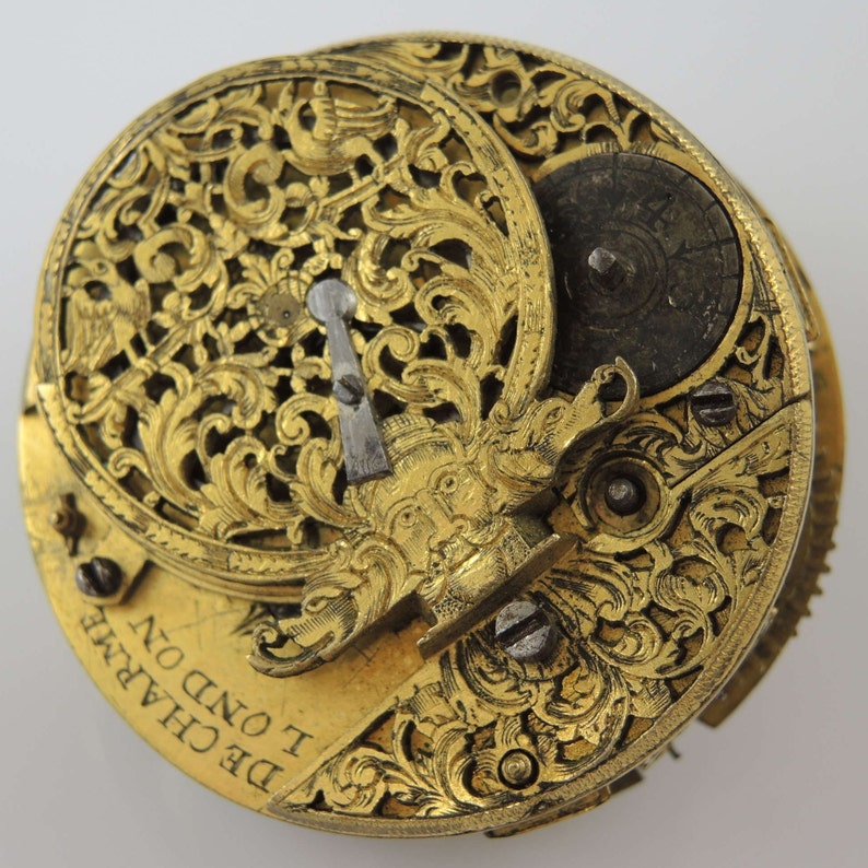 May include: A gold pocket watch with intricate floral engravings and a detailed face. The watch face has a small, round window and the text "DE CHAINE I OND ON" is visible on the bottom.