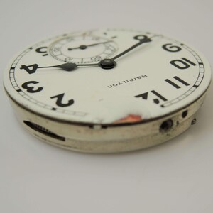 May include: A vintage watch face with a white background, black numerals, and black hands. The watch face has a subdial and the word "HAMILTON" printed on it. The watch face is in a silver-coloured casing.