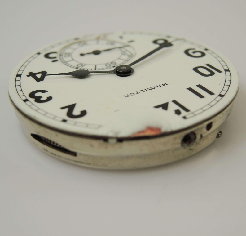 May include: A vintage watch face with a white background, black numerals, and black hands. The watch face has a subdial and the word "HAMILTON" printed on it. The watch face is in a silver-coloured casing.