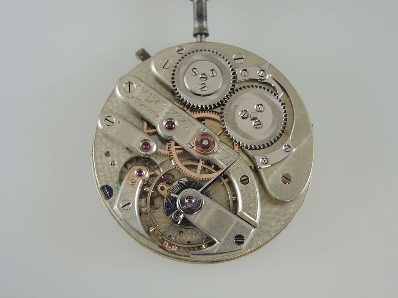 May include: Close-up of a silver watch movement with intricate gears and a rose gold finish. The movement is visible through a clear case back.
