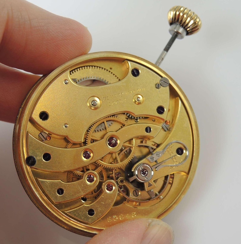 May include: Close-up of a gold-toned pocket watch movement with intricate gears and ruby jewels. The watch is marked "Swiss Made" and has a serial number.