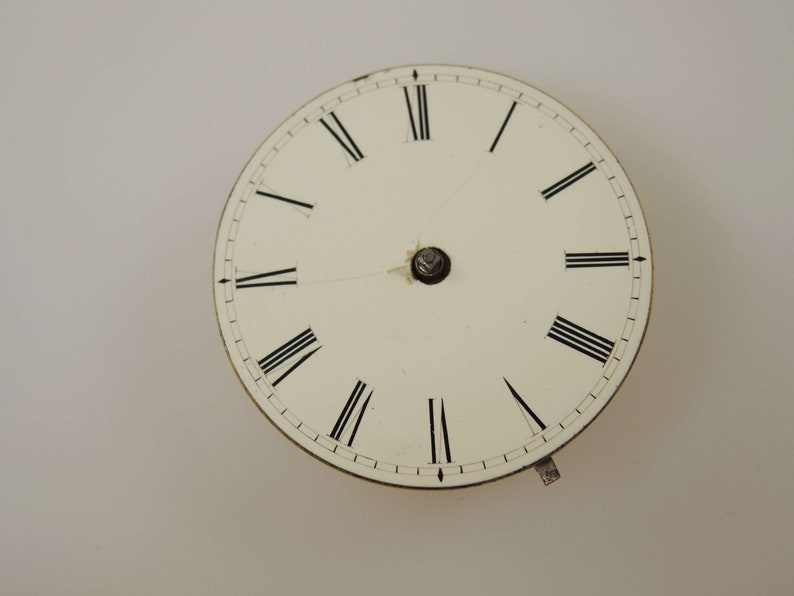 May include: A white clock face with black Roman numeral hour markers and black minute markers. The clock face is circular and has a silver center.
