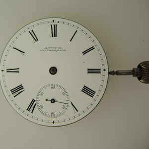 May include: Close-up of a vintage pocket watch face. The white face features black Roman numerals, a smaller seconds dial, and the text "A.W.W. Co., Waltham, Mass." A gold-colored winding stem and crown are attached.