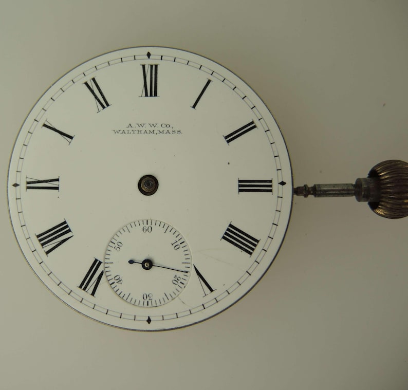 May include: Close-up of a vintage pocket watch face. The white face features black Roman numerals, a smaller seconds dial, and the text "A.W.W. Co., Waltham, Mass." A gold-colored winding stem and crown are attached.