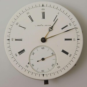 May include: A classic pocket watch face with a white dial, black Roman numerals, and a sub-dial for seconds. The watch features black hands and the name "CHAS. JACOB" is printed on the face.