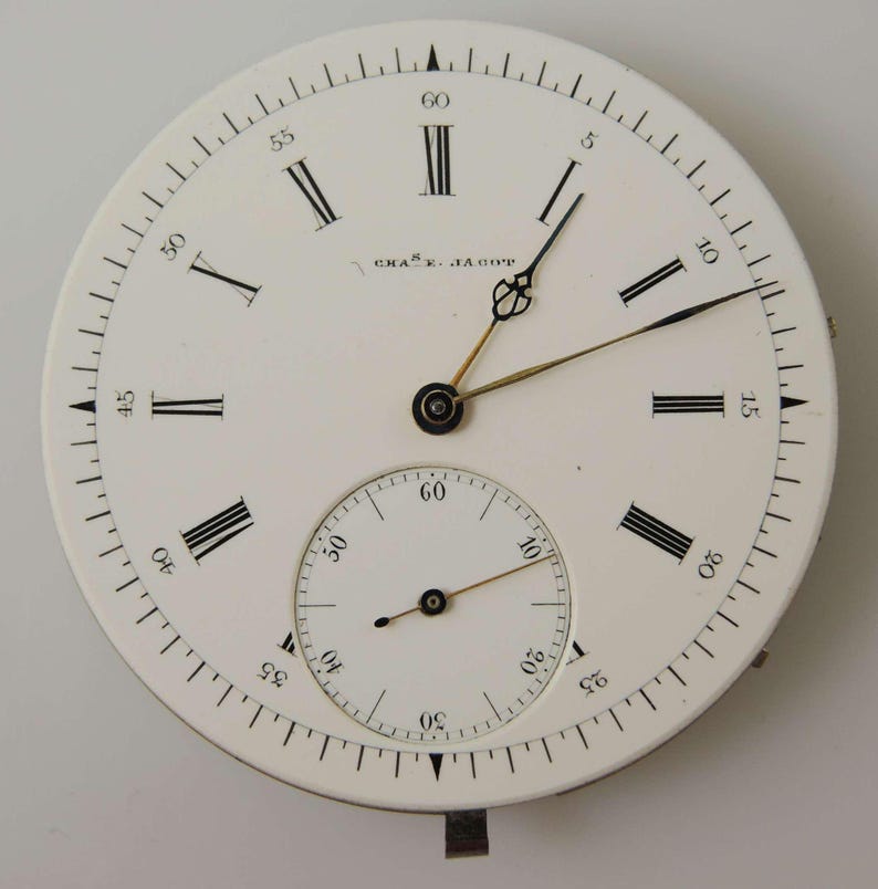 May include: A classic pocket watch face with a white dial, black Roman numerals, and a sub-dial for seconds. The watch features black hands and the name "CHAS. JACOB" is printed on the face.
