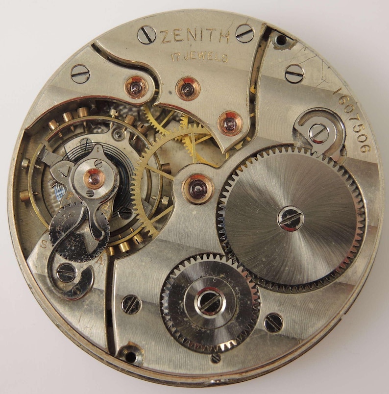 May include: Close-up of a Zenith watch movement, showcasing intricate gears, springs, and jewels. The silver-toned metal components are arranged in a circular pattern, with the word "ZENITH" and "17 JEWELS" visible. The serial number "1607506" is also present.