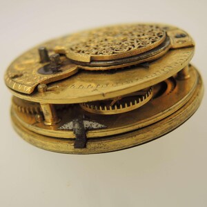 Puede incluir: Primer plano del mecanismo de un reloj de bolsillo antiguo. La imagen muestra los intrincados engranajes y una placa decorativa de filigrana dentro del reloj. Los componentes met&aacute;licos tienen un tono dorado, posiblemente lat&oacute;n u oro. El reloj est&aacute; parcialmente desmontado.
