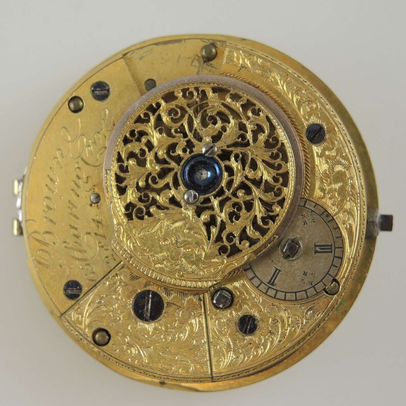 Puede incluir: Un reloj de bolsillo de oro con un dise&ntilde;o intrincado grabado. La esfera del reloj es visible y presenta n&uacute;meros romanos. La inscripci&oacute;n "John Brown London" es visible en la esfera del reloj.