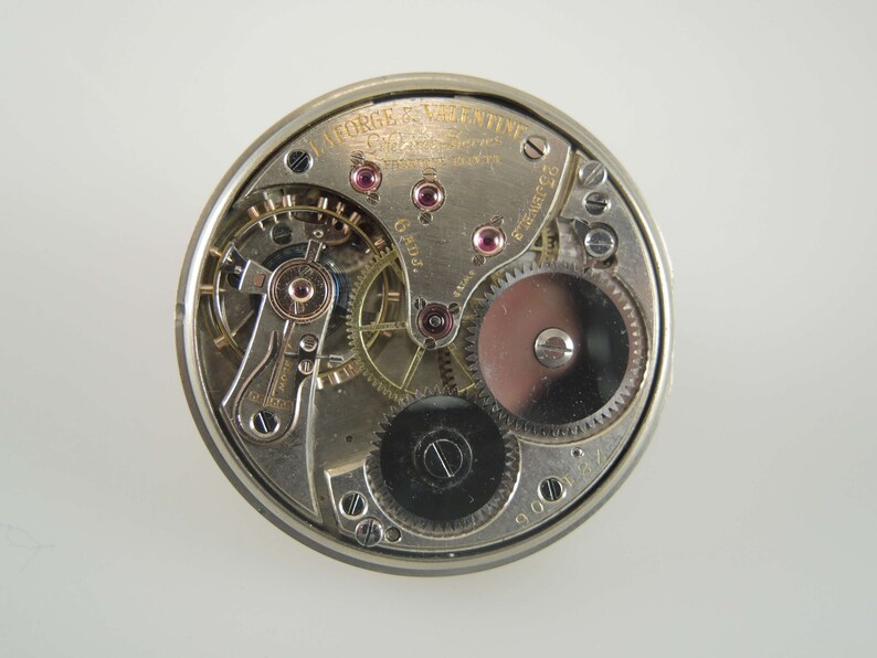 K&ouml;nnte beinhalten: Nahaufnahme eines silbernen Taschenuhrwerks mit der Aufschrift "W. L. Forge & Valentine Chicago, Ill. 6 Adj. Patent Applied For" und der Nummer "900127".