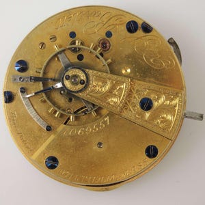 May include: Close-up of a gold pocket watch movement with a serial number 2069557. The watch has a decorative engraved plate and blue screws.