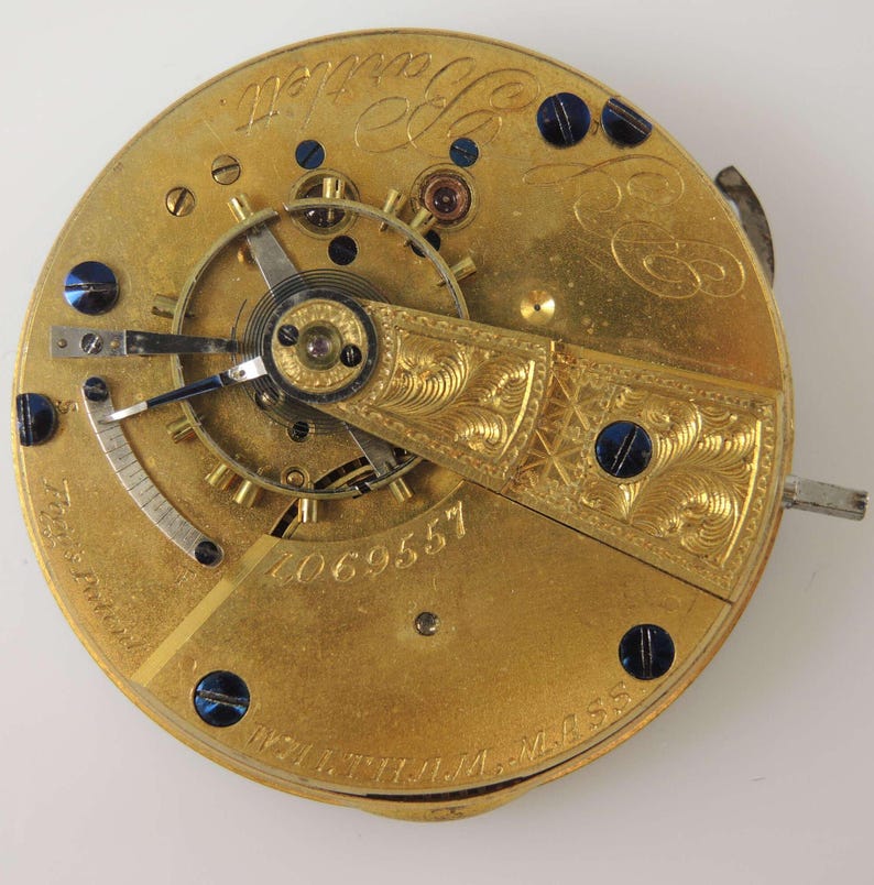 May include: Close-up of a gold pocket watch movement with a serial number 2069557. The watch has a decorative engraved plate and blue screws.