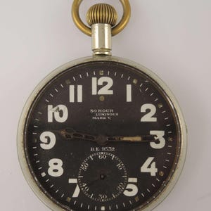 Op de afbeelding: Een vintage zakhorloge met een zwarte wijzerplaat en witte cijfers. Het horloge heeft een zilverkleurige kast en een goudkleurige ring aan de bovenkant. De tekst "30 HOUR LUMINOUS MARK V" is zichtbaar. Het horloge heeft ook een kleinere wijzerplaat.