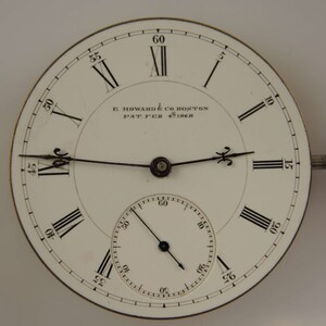 Puede incluir: Una esfera de reloj de bolsillo blanca con marcadores de hora en n&uacute;meros romanos negros y agujas negras. La esfera del reloj tiene un c&iacute;rculo conc&eacute;ntrico m&aacute;s peque&ntilde;o con marcas negras para los segundos. La esfera del reloj est&aacute; inscrita con "E. HOWARD & CO BOSTON PAT. FEB 4th 1868".