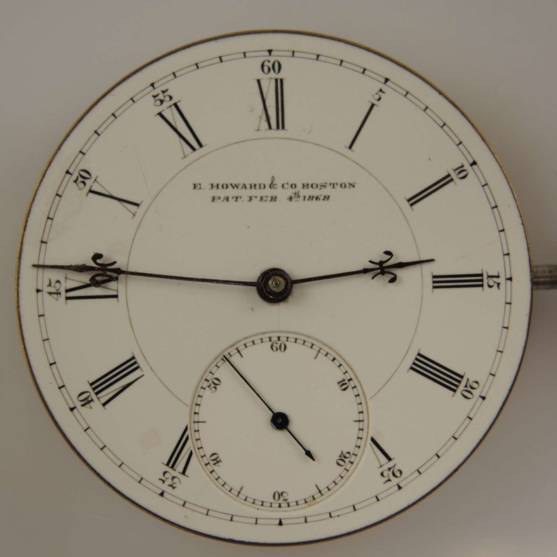 Puede incluir: Una esfera de reloj de bolsillo blanca con marcadores de hora en n&uacute;meros romanos negros y agujas negras. La esfera del reloj tiene un c&iacute;rculo conc&eacute;ntrico m&aacute;s peque&ntilde;o con marcas negras para los segundos. La esfera del reloj est&aacute; inscrita con "E. HOWARD & CO BOSTON PAT. FEB 4th 1868".