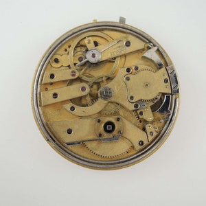 Op de afbeelding: Close-up van een goudkleurig zakhorloge-uurwerk met ingewikkelde tandwielen en hendels. Het horloge is gedemonteerd en de interne werking is zichtbaar.