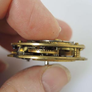 May include: Close-up of a vintage watch movement with visible brass gears and components. The intricate mechanism is held between two fingers, showcasing the detailed craftsmanship of the timepiece. The gears are a warm gold color.