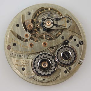 May include: Close-up of a vintage Hamilton pocket watch movement. The silver-toned mechanism features intricate gears, screws, and jewels. The watch face is marked with "Hamilton, Lancaster, PA," and "Adjusted 5 Positions, 23 Jewels."