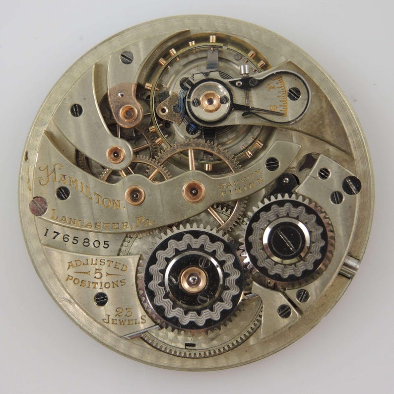 May include: Close-up of a vintage Hamilton pocket watch movement. The silver-toned mechanism features intricate gears, screws, and jewels. The watch face is marked with "Hamilton, Lancaster, PA," and "Adjusted 5 Positions, 23 Jewels."