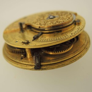May include: Close-up of a vintage brass pocket watch mechanism. The watch features intricate gears, dials, and hands, with the word "East" inscribed on the dial. The watch has a golden hue and is set against a white background.