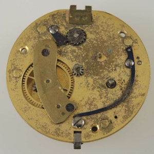 Puede incluir: Primer plano de un movimiento de reloj dorado con engranajes intrincados y una palanca de metal azul. El movimiento se coloca sobre un fondo liso y claro.
