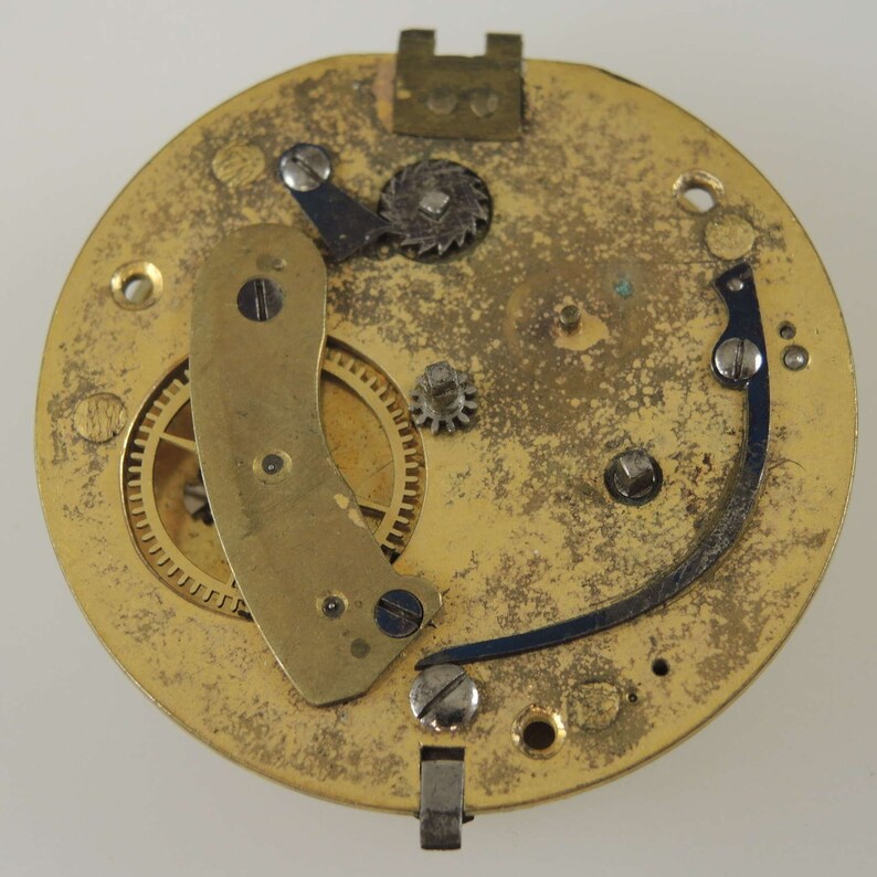 Puede incluir: Primer plano de un movimiento de reloj dorado con engranajes intrincados y una palanca de metal azul. El movimiento se coloca sobre un fondo liso y claro.