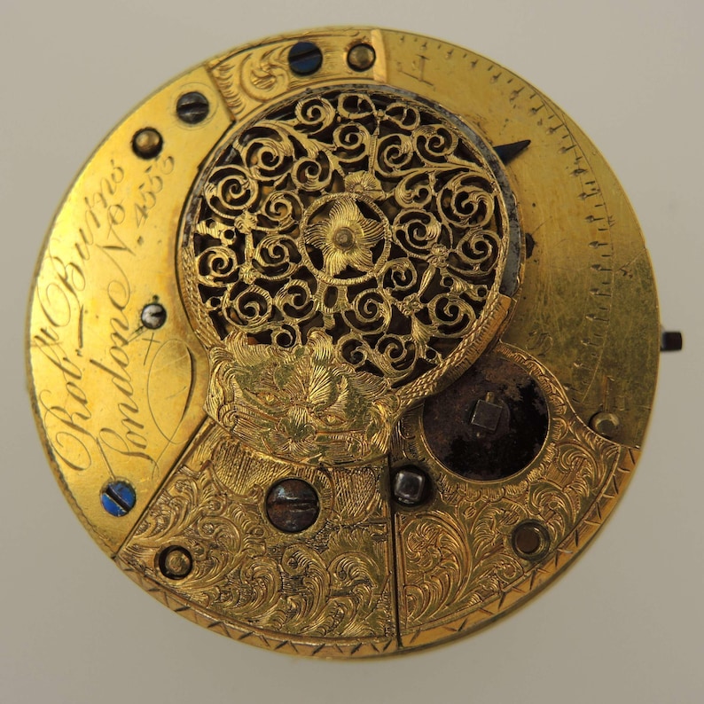 Puede incluir: Primer plano de un movimiento de reloj de oro vintage. El mecanismo del reloj presenta intrincados grabados florales y una cubierta de filigrana decorativa. El texto "Robt. Burgess London" est&aacute; inscrito en la superficie dorada. Est&eacute;tica cl&aacute;sica y antigua.