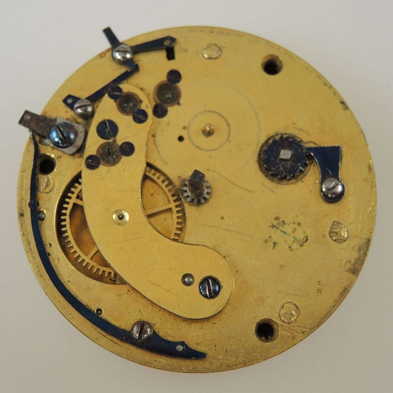 Puede incluir: Primer plano de un movimiento de reloj de color dorado con engranajes intrincados y un marco de metal negro. El movimiento se coloca sobre un fondo blanco.