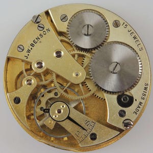 Puede incluir: Primer plano de un movimiento de reloj de color dorado con engranajes intrincados y una placa plateada que dice "15 Jewels Swiss Made". El movimiento tambi&eacute;n tiene la inscripci&oacute;n "J.W. Benson" en la parte superior izquierda.