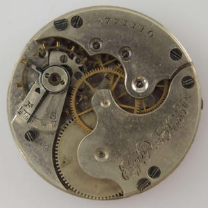 May include: Close-up of a vintage watch movement, showcasing intricate gears, screws, and a balance wheel. The metal components are silver-toned with gold-colored gears. The text "Elgin Nat'l Watch Co." is visible.