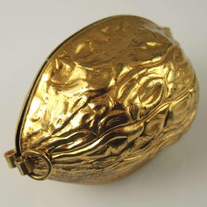 May include: A gold-colored metal container shaped like a walnut shell. The object features a highly textured, irregular surface mimicking the natural ridges of a nut. A small, circular metal ring is attached to one end of the hinged shell.