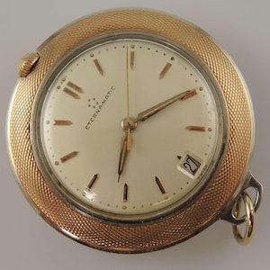 Puede incluir: Un reloj de pulsera Eterna-Matic vintage con caja dorada y esfera color crema. El reloj presenta agujas e indicadores dorados, una pantalla de fecha y el texto "ETERNA-MATIC". El reloj tiene un bisel texturizado y una pequeña corona.