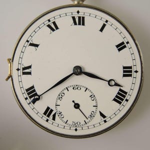 Puede incluir: Primer plano de un reloj de bolsillo vintage con esfera blanca y n&uacute;meros romanos negros. El reloj tiene una esfera secundaria m&aacute;s peque&ntilde;a para los segundos y agujas negras. El reloj est&aacute; en una caja de metal plateado.