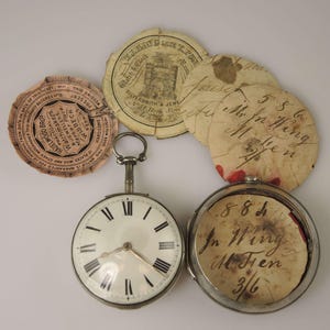 Könnte beinhalten: Eine silberne Taschenuhr mit römischen Ziffern auf dem Zifferblatt. Die Uhr ist geöffnet und die Innenseite des Gehäuses trägt eine handschriftliche Aufschrift mit dem Text "884 In Wing M Fren 3/6". Es gibt vier kleine Papierkreise mit handschriftlichem Text darauf.