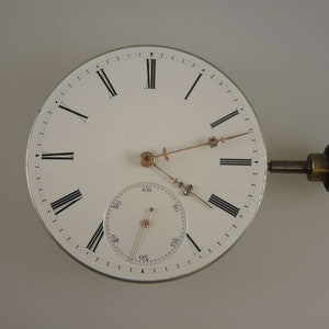 May include: A white pocket watch with Roman numeral hour markers and a smaller sub-dial for seconds. The watch has a gold-colored winding key and hands.